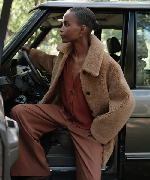 A woman with a pulled-back hairstyle sits in the open car doorway, wearing the Shearling Aurora Coat over a rust-red cardigan and wide-leg brown pants, gazing calmly to the side.