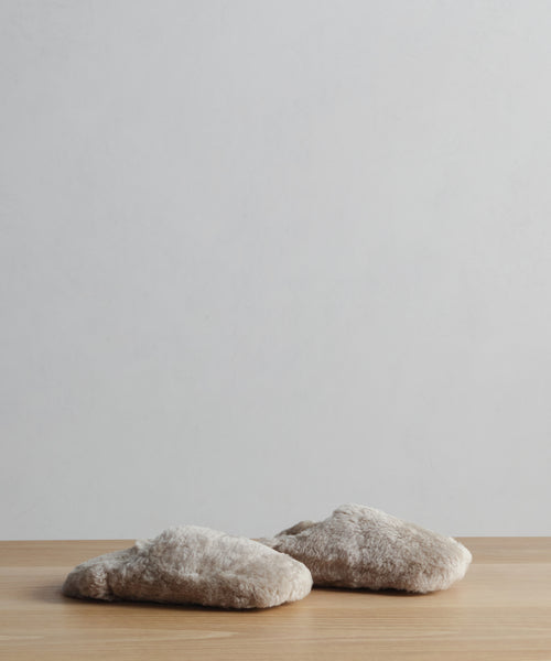 The Shearling Moroccan Slipper, a beige and fluffy pair with a suede outsole, rests on a wooden surface against a plain, light-colored wall.
