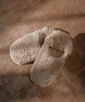 A pair of Shearling Moroccan Slippers in beige with fluffy uppers and soft suede outsoles rest on a textured brown surface, gentle shadows falling across them.