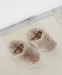 A pair of Shearling Moroccan Slippers in light brown with a soft, fuzzy texture and suede outsole sit on a cream woven rug beside a marble surface.
