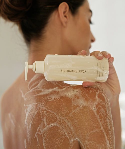 A woman bathes, showcasing her shoulder and holding the Silk Body Duo, highlighting its skin-soothing, hydrating formula and clear label.