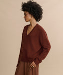Woman with curly hair wears the Simone V-Neck in rust, paired with a brown skirt, standing sideways against a beige background and looking forward with a relaxed expression—a luxurious layer for any occasion.