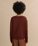 Seen from behind, a person with curly hair wears the Simone V-Neck sweater and brown pants—a loose-fitting luxury layer styled against a plain beige background.