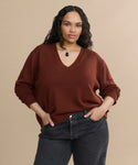 A woman with long, wavy hair poses confidently against a beige background wearing the rust-colored Simone V-Neck sweater, dark jeans, and a black pendant necklace. Her hands are in her pockets.