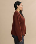 Woman with long dark hair wears the Simone V-Neck sweater in rust, paired with dark jeans, standing in profile against a beige background—a versatile cotton-merino luxury layer for any occasion.