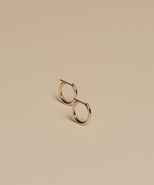 The Small Eloise Hoops, crafted from 14k gold by Jenni Kayne, are displayed slightly apart on a plain beige surface with soft lighting and minimal shadows.