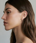 A woman with long brown hair is shown in profile against a plain background, wearing Small Eloise Hoops—delicate 14k gold earrings. She has clear skin and a neutral expression.
