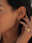 Close-up of a person’s ear wearing Small Eloise Hoops by Jenni Kayne. Their manicured hand touches the ear, highlighting a chunky gold ring—an elegant mix of fine jewelry against freckled skin.