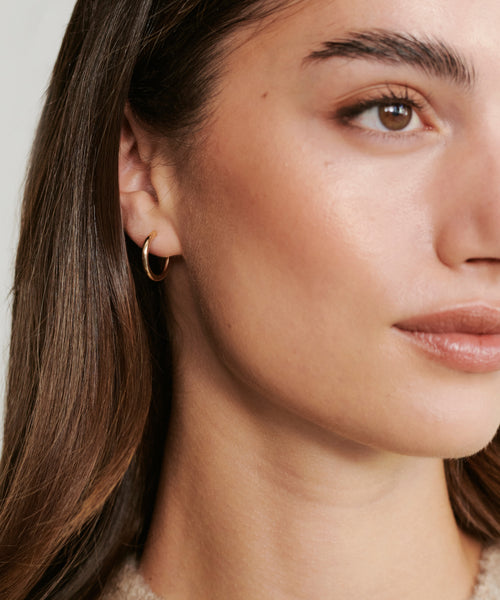 Close-up of a woman’s face with smooth skin, natural makeup, and straight brown hair. She wears a Small Eloise Hoop earring by Jenni Kayne in her left ear and looks slightly to the right.