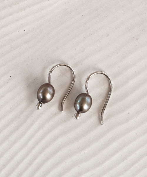 The Mermaid Earrings are sterling silver hooks, each adorned with a single dark gray freshwater pearl, shown on a textured, light-colored surface with subtle wavy lines.