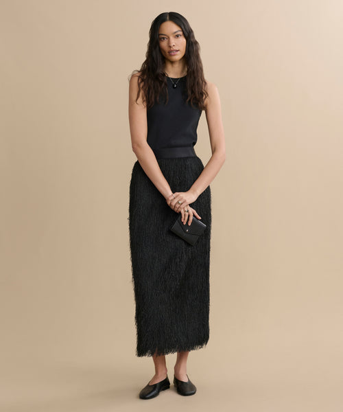 A woman with long wavy hair wears a sleeveless black top and the Sterling Skirt, holding a small black clutch and wearing black flats, standing against a plain beige background.