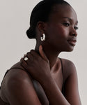A woman with dark skin and slicked-back hair wears the Large Donut Hoops in sterling silver, a chunky silver ring, and a brown camisole as she gazes thoughtfully sideways, her hand resting on her neck.