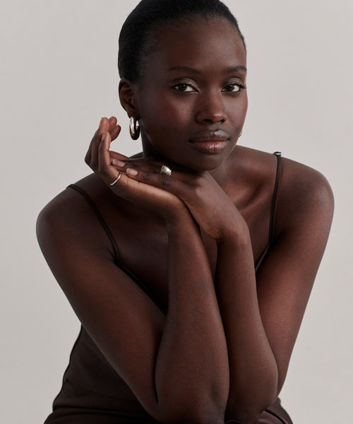 A woman with dark skin and short hair, wearing a sleeveless brown top and Large Donut Hoops, rests her chin on her hands and looks calmly at the camera against a plain light background.