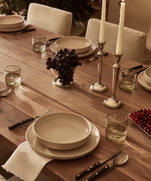 A wooden dining table set with Pacific Soup Bowl, beige plates, silverware, glasses of light drink, a bowl of grapes, a tray of cherries, and two tall lit candles in silver holders. White napkins are folded under the stoneware dinner plate.