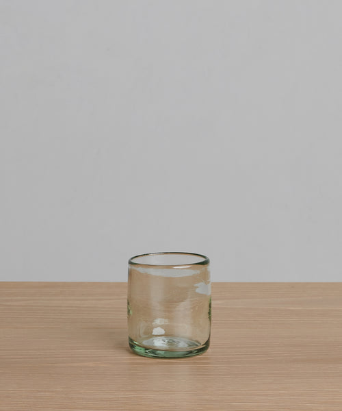 The Straight Rock Glass, handblown from recycled glass with a light green tint, rests on a pale wooden surface against a plain light gray background—a perfect sustainable glassware choice.