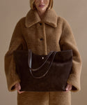 A person in a tan, oversized faux fur coat holds the Suede Agnes Tote, a large dark brown suede bag with thin handles, against a plain beige background. Their face is partially visible, cropped above the mouth.