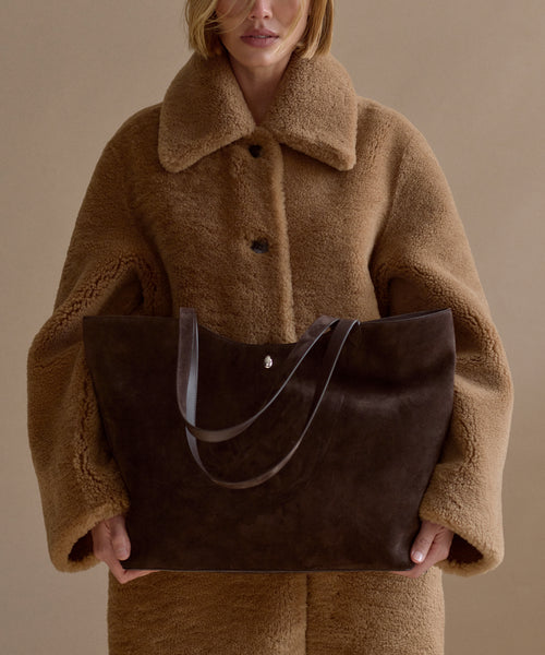 A person in a tan, oversized faux fur coat holds the Suede Agnes Tote, a large dark brown suede bag with thin handles, against a plain beige background. Their face is partially visible, cropped above the mouth.