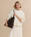 A woman with short blonde hair, dressed in a cream cable-knit sweater and light skirt, holds the Suede Agnes Tote in dark brown, standing against a beige background.