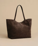 The Suede Agnes Tote is a large dark brown suede bag with slim black leather shoulder straps, shown on a plain beige background. Ideal as a chic laptop bag, it includes a small round metal button near the top center.