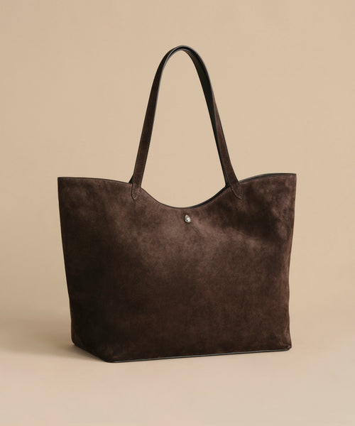 The Suede Agnes Tote is a large dark brown suede bag with slim black leather shoulder straps, shown on a plain beige background. Ideal as a chic laptop bag, it includes a small round metal button near the top center.