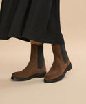 A person in a long black skirt wears the Suede Alden Boot—brown suede Chelsea boots with black elastic side panels and a weather-friendly rubber sole—standing on a light beige background.