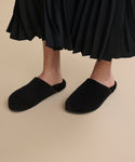 A person wearing Suede Ashby Clogs featuring a black slip-on design and shearling-lined footbed stands on a beige floor, paired with a black pleated skirt.