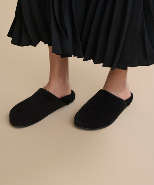 A person wearing Suede Ashby Clogs featuring a black slip-on design and shearling-lined footbed stands on a beige floor, paired with a black pleated skirt.