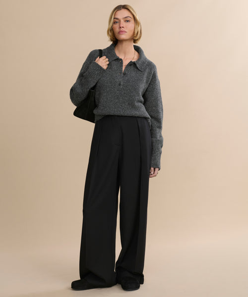 A person stands against a beige background wearing a gray pullover, loose black trousers, black shoes, and a black bag, with short wavy hair completing the look—finished with classic Suede Ashby Clog footwear.