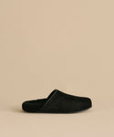 The Suede Ashby Clog, a black slip-on with a fuzzy shearling-lined footbed and closed toe, is shown against a beige background.