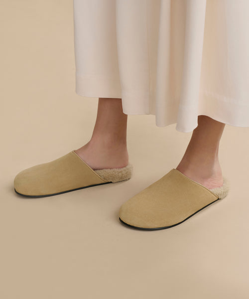 A person wearing a long cream skirt and Suede Ashby Clog slip-ons with a shearling-lined footbed, standing on a light tan floor.