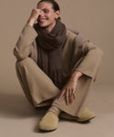 A person sits cross-legged on the floor, smiling with one hand on their face. They wear beige loungewear, a brown scarf, gold jewelry, and the Suede Ashby Clog—featuring a cozy shearling-lined footbed—against a beige background.