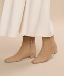 A person wearing a long, cream-colored skirt and Suede Carmine Boot low-heeled booties stands on a light beige surface.