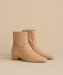 A pair of Suede Carmine Boot taupe suede booties with low block heels and pointed toes, displayed on a beige surface against a matching beige background.