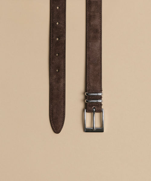The Suede Danner Belt in brown, with a silver rectangular buckle, is laid flat on a beige background. This belt features multiple adjustment holes and two metal loops near the buckle.