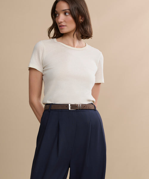 A person wears a short-sleeve cream t-shirt tucked into high-waisted navy trousers with the Suede Danner Belt, standing against a beige background and looking to the side.