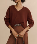 Woman wearing a loose, rust V-neck sweater tucked into high-waisted brown trousers with the Suede Danner Belt, holding a black strap against a neutral background.