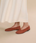 Wearing a long, cream skirt and rust-colored Suede Elodie Flats, a person stands on a beige floor, creating a graceful and feminine silhouette.