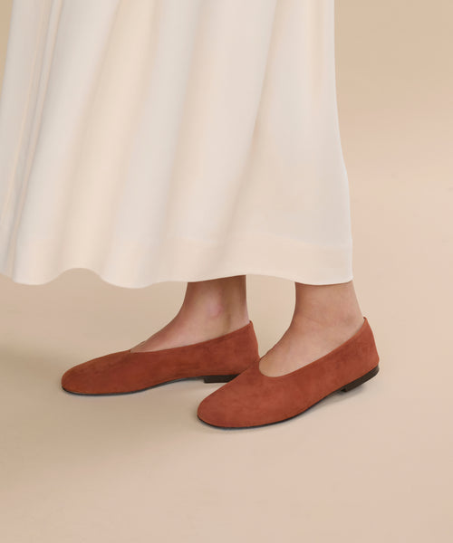 Wearing a long, cream skirt and rust-colored Suede Elodie Flats, a person stands on a beige floor, creating a graceful and feminine silhouette.