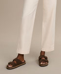 Only the lower legs and feet are visible as someone models the Suede Hutton Slide Sandal in brown suede with push-button detailing, paired with off-white pants against a neutral background—a perfect warm-weather shoe.