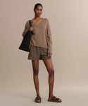 A woman stands against a beige background in a brown V-neck sweater, brown shorts, hoop earrings, a black textured shoulder bag, and completes her look with chic Suede Hutton Slide Sandals.