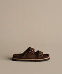 The Suede Hutton Slide Sandal, shown in profile against a beige background, features brown suede, two adjustable buckle straps, and a flat sole—perfect for warm-weather style.