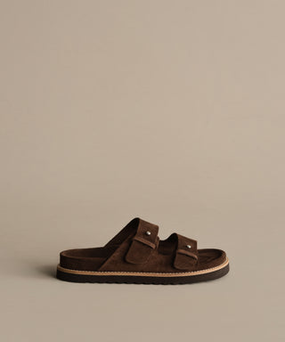 The Suede Hutton Slide Sandal, shown in profile against a beige background, features brown suede, two adjustable buckle straps, and a flat sole—perfect for warm-weather style.