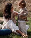 A woman in Suede Hutton Slide Sandals smiles at a young child as they enjoy time together outdoors in neutral sweaters and light pants among tall grasses—a perfect moment for their favorite warm-weather shoes.