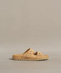 The Suede Hutton Slide Sandal, a tan suede slide with two buckle straps and a ridged sole, is shown against a plain beige background.