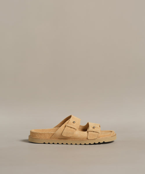 The Suede Hutton Slide Sandal, a tan suede slide with two buckle straps and a ridged sole, is shown against a plain beige background.