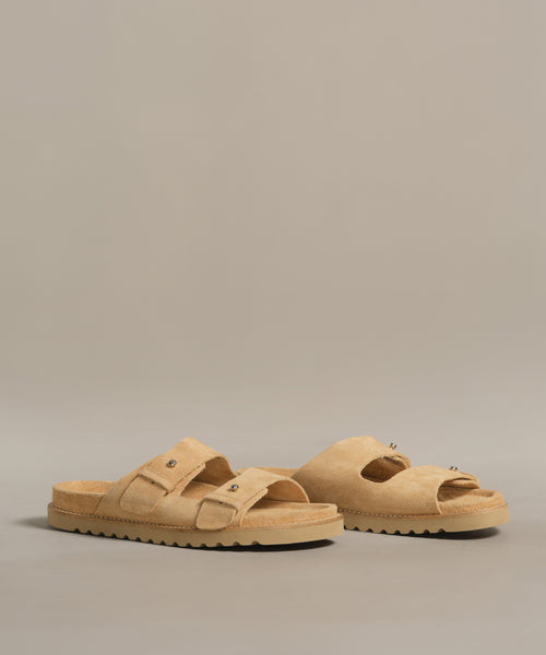 The Suede Hutton Slide Sandal, with tan suede, two adjustable buckle straps, and textured rubber soles, is shown on a gray background—an effortless and stylish option for warm weather.