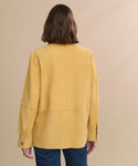 A person with shoulder-length brown hair is shown from the back, wearing a loose-fitting Suede Lennox Jacket in a honey shade and blue jeans, standing against a neutral beige background.