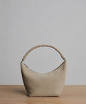 The Medium Crescent Bag in beige suede features a curved silhouette and twisted handle. Displayed on wood with a light gray background. Crafted in Italy.