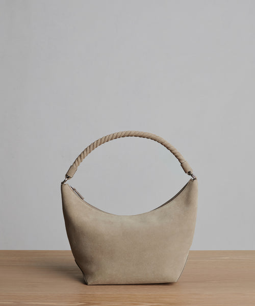The Medium Crescent Bag in beige suede features a curved silhouette and twisted handle. Displayed on wood with a light gray background. Crafted in Italy.