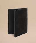 The Suede Passport Holder in black stands upright against a plain beige background, highlighting its soft texture and minimalist design—perfect for organizing your travel essentials.